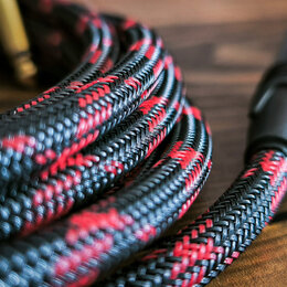 Detailní záběr na svinutý kabel v textilním opletení. Kabel má černou barvu s červenými vzory a je zakončen černým plastovým konektorem. Povrch oplet…