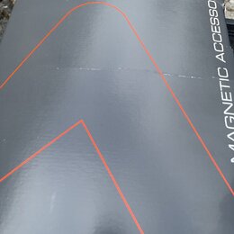 Detailní záběr na šedý povrch s nápisem MAGNETIC ACCESSORIES. Na povrchu jsou vidět tenké červené linky tvořící geometrické tvary. Materiál vykazuje…