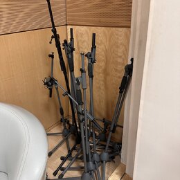 Skupina několika šedých mikrofonních stojanů s trojnožkami, opřených o stěnu v rohu místnosti. Stojany mají kovovou konstrukci a černé plastové úchyt…