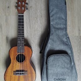 Dřevěná ukulele v přirozené hnědé barvě s černým hmatníkem a čtyřmi strunami. Na hlavě nástroje i na spodní části šedého textilního obalu je uveden n…