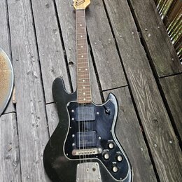 Černá elektrická kytara s dřevěným hmatníkem a hlavou, položená na dřevěné terase. Nástroj má černý pickguard a dva snímače typu humbucker. Povrch tě…