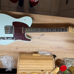 Světle modrá elektrická kytara typu Telecaster s červeně hnědým vzorovaným pickguardem a hnedým hmatníkem. Na hlavě kytary je vidět nápis Fender TELE…