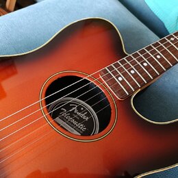 Akustická kytara s barevným přechodem od oranžové k tmavě hnědé (sunburst). Uvnitř ozvučovacího otvoru je štítek s nápisy Fender, Telecoustic, ELECTR…