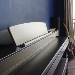 Černý digitální piano s doprovodnou stoličkou. Na horní části korpusu je vidět vybledlý nápis YAMAHA. Na nástroji je umístěn bílý notový stojan. V po…