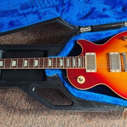 Elektrická kytara s trupem v červeno-žlutém přechodovém odstínu (sunburst) umístěná v černém tvrdém kufru s modrým polstrováním. Na hlavě hmatníku je…