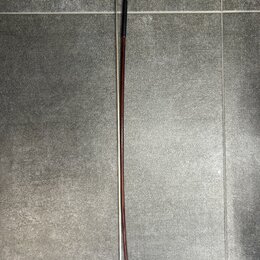 Fotografie znázorňuje smyčec z tmavého dřeva s kovovými a plastovými prvky. Na horní části je vidět černý plastový šroubovací mechanismus s malým zla…