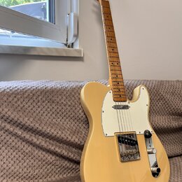 Elektrická kytara typu Telecaster v krémové barvě s bílým pickguardem. Na hlavě hmatníku je viditelné logo Fender. Nástroj je vybaven stříbrným moste…