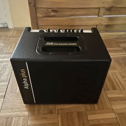Černý kombozesilovač s texturovaným povrchem a integrovanými madly. Na horní části je nápis AER The Acoustic People a na boční straně vertikální bílý…