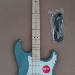 Modrá elektrická kytara typu Stratocaster se světle hnědým hmatníkem a bílým pickguardem. Na hlavě je černý nápis Squier a menší text by Fender. Na t…