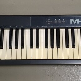 Černá klaviatura s bílými a černými klávesami. Na horní liště je vlevo nápis KEYSTATION61 a vpravo M-AUDIO. V levé části jsou umístěny posuvníky, dva…