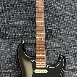Elektrická kytara s černým bodies, hnědým hmatníkem a světle hnědou hlavou. Na hlavě je nápis Fender Stratocaster. Kytara má tři bílé snímače, tři bí…