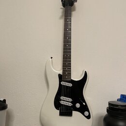 Bílá elektrická kytara s černým hmatníkem a černým pickguardem. Na hlavě je bílý nápis Squier STRATOCASTER. Nástroj má tři snímače, jeden ovládací kn…