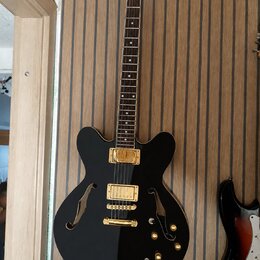 Černá polodubová elektrická kytara s gold hardwarem a hnědým hmatníkem. Nástroj má dva humbuckery, most s laděním strun a tři ovládací knoflíky v zla…