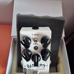 Bílý efekt pedál s nápisy BAS TARD a LOW END DISTORTION v bílé krabičce s šedou pěnovou vložkou. Pedál má čtyři černé oválné ovládací knoflíky, jeden…