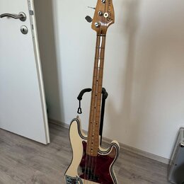 Elektrická basová kytara v krémové barvě s tmavě červeným pickguardem, umístěná na černém stojanu. Na hlavě nástroje je nápis „EMELIA PRECISION BASS“…