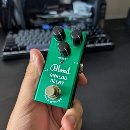 Zelený efekt pedál s kovovým povrchem. Na přední straně je bílý nápis „Blond ANALOG DELAY“ a v dolní části text „TRUE BYPASS“. Pedál má tři černé ovl…