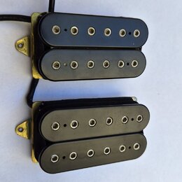 Dvě černé elektrické kytarové snímače typu humbucker s kovovými zlatými rámečky. Každý snímač má dvě řady kulatých pólů. Povrch plastových krytů je h…