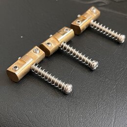 Tři kovové mostkové sedla v mosazné barvě s pružinami. Každé sedlo obsahuje dva malé imbusové šroubky pro ladění výšky. Povrch je hladký, bez viditel…