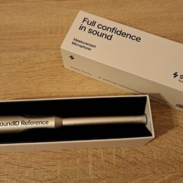 Bílý papírový obal a otevřená krabice s měřicím mikrofonem stříbrné barvy. Na obalu je nápis Full confidence in sound, Measurement Microphone a logo…