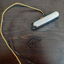 Na tmavé dřevěné desce leží černý magnetický snímač s chromovaným krytem a dvěma přilehlými šrouby. Ze snímače vycházejí dva dlouhé vodiče, jeden v č…