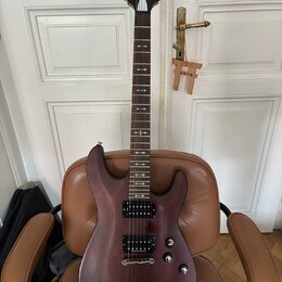 Elektrická kytara s tmavě hnědým, polomatným povrchem těla a hnedým hmatníkem. Na hlavě je bílý nápis Schecter Diamond Series. Nástroj má dva humbuck…
