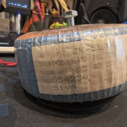 Fotografie znázorňuje elektrický transformátor s modrým lakovaným jádrem, omotaný průhlednou fólií. Na boku je nalepený papírový štítek s ručně psaný…