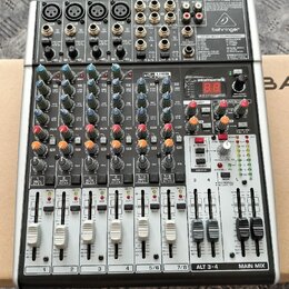 Stříbrný mixážní pult značky behringer s černým šasi. Na horním panelu je nápis 2-TRACK/USB a modelové označení XENYX X1204USB. Zařízení obsahuje něk…