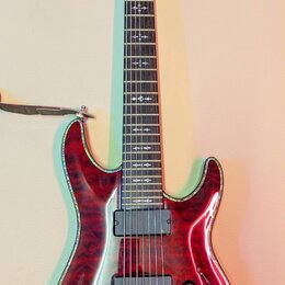 Elektrická kytara s červeným lakovaným tělem s výraznou strukturou dřeva a lemováním. Na hlavě je nápis Schecter. Nástroj má dva humbuckery, tři ovál…