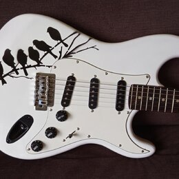 Bílá elektrická kytara s tvarem typu Stratocaster a bílým pickguardem. Na horní části korpusu je černý grafický motiv větve s pěti ptáky. Nástroj má…