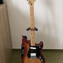 Elektrická kytara s tělem v červenohnědém sunburst provedení a světlým javorovým hmatníkem. Na hlavě je viditelné logo Fender a na popruhu nápis Fend…