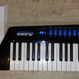 Černý MIDI kontrolér s klávesnicí a modrým podsvícením displeje a indikátorů. Na přístroji je bílý nápis ALESIS VORTEX WIRELESS 2. Vedle nástroje lež…