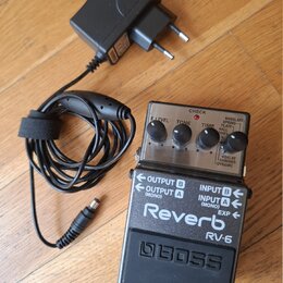 Na dřevěné podlaze leží efekt pedál Boss RV-6 Reverb v černé barvě s kovovým horním panelem a černý síťový adaptér. Na pedálu jsou nápisy „Reverb“, „…
