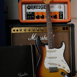Elektrická kytara typu Stratocaster s barevným přechodem od žluté přes oranžovou k hnědé a bílým pickguardem. Na hlavě krku je viditelné logo Fender.…