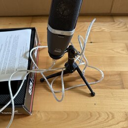 Stříbrný mikrofon s černou mřížkou umístěný na černém tripodním stativu. Na těle mikrofonu jsou nápisy MONITOR, 22040955 a ABx555. Vedle mikrofonu je…