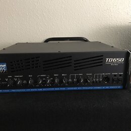Černý basový zesilovač s kovovým šasi a madlem. Na předním panelu je vlevo logo EBS a vpravo nápis TD650 Professional Tube Definition Bass Head. Dole…