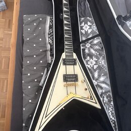 Bílá elektrická kytara s tvarem V v černém obalu. Na hlavě hmatníku je nápis Jackson. Kytara má dva humbuckery, zlatý hardware a bílý lem kolem těla.…