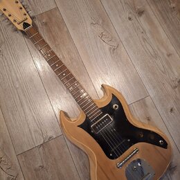 Elektrická kytara s přírodním dřevěným povrchem a černým pickguardem. Na hlavě hmatníku je vidět nápis "Epiphone". Nástroj vykazuje známky opotřebení…