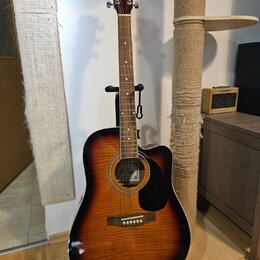 Akustická kytara s cutawayem v tmavě hnědé a oranžové barvě (sunburst) umístěná na černém stojanu. Na hlavě hmatníku je nápis M. Polak a v otvoru rez…