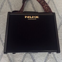 Černý akustický kombozesilovač s texturovaným povrchem a čelní stofovou mřížkou. V horní části je umístěno zlaté logo NUX a pod ním nápis ACOUSTIC. K…