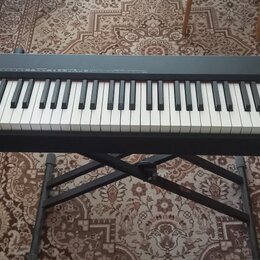 Černý klavírní kontrolér značky Roland, model A-88. Nástroj je umístěn na černém X-stojanu. Na levé straně jsou ovládací prvky, knofíky a tlačítka. P…