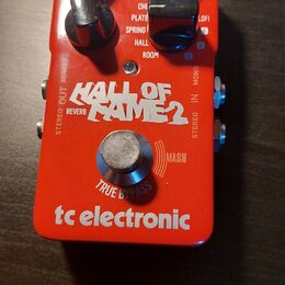 Červený efekt pedál s nápisem HALL OF FAME 2 REVERB a logem tc electronic. Na horní části jsou tři černé otočné ovladače a přepínače s popisky PRE-DE…