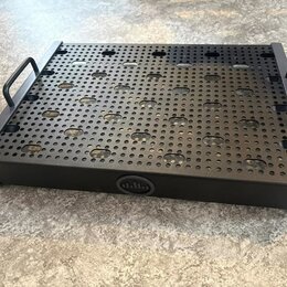 Černý kovový pedalboard s perforovaným vrchem a dvěma bočními madly. Na přední hraně je umístěn kulatý emblem s logem znázorňujícím stylizované sloup…