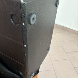 Detail černé reproduktorové bedny s texturovaným povrchem. Na spodní straně jsou vidět čtyři gumové nožičky a na boku kulatý úchop. Hrany bedny vykaz…
