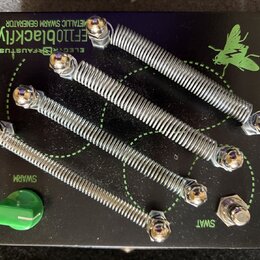 Černý efekt pedál s nápisy v zelené barvě: „EF110blackfly“, „METALLIC SWARM GENERATOR“ a „ELEGY FAUSTUS“. Na povrchu jsou čtyři kovové pružiny připev…