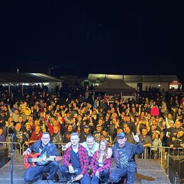 Fotografie zachycuje skupinu lidí před pódiem v noci. V popředí klečí tři muži a jedna žena, přičemž muž vlevo drží červenou elektrickou basovou kyta…