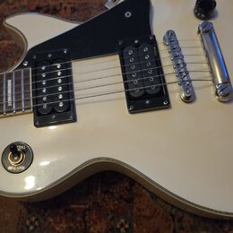Bílá elektrická kytara s černým pickguardem a dvěma černými humbuckery. Na přepínači tónu jsou viditelné nápisy TREBLE a RHYTHM. Nástroj má chromovan…