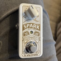 Bílý efekt pedál Spark Mini Booster značky tc electronic. Na přední straně je bílý oválný ovládací knoflík a stomečový spínač True Bypass. Jsou zde n…