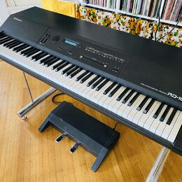 Černý digitální klavír na kovovém stojanu s přiloženým černým pedálem. Na horní části je nápis Roland a vpravo u kláves označení ROLAND DIGITAL PIANO…
