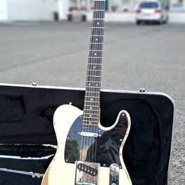 Elektrická kytara typu Telecaster v krémové barvě s černým pickguardem, umístěná v otevřeném černém tvrdém kufru. Na hlavě hmatníku je viditelné logo…