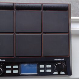 Černý elektronický bicí pad s devíti čtvercovými polštáři v mřížce 3x3. Na spodním panelu jsou nápisy Millenium a NonaPad, malý displej, několik tlač…